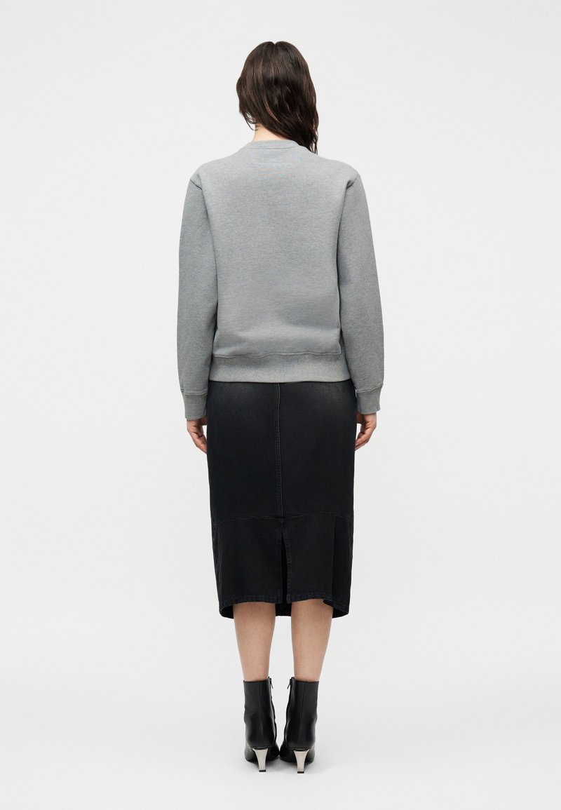 Femme aux longs cheveux foncés portant un sweat-shirt gris, une jupe midi noire avec une fente au dos, et des bottines noires à talons argentés, debout de dos.