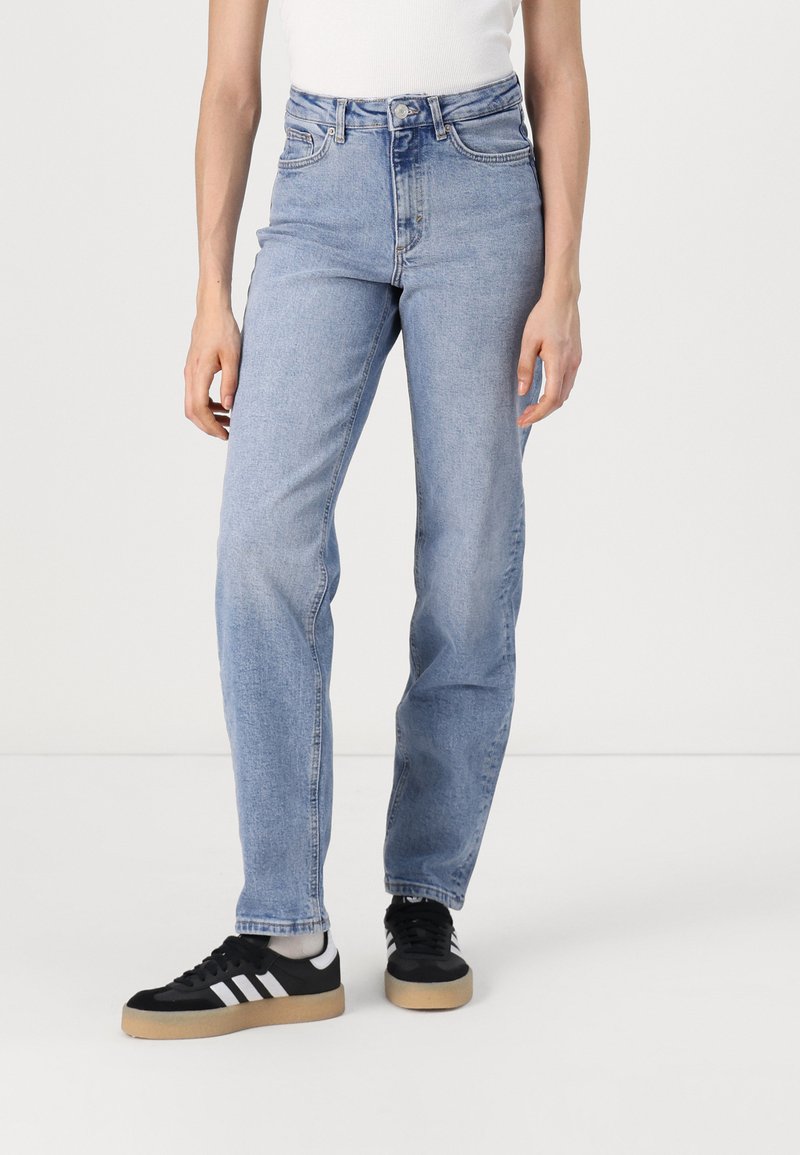Jeans de tiro alto en azul claro, hechos de denim, con piernas rectas y un ligero desgastado, combinados con zapatillas negras y una camiseta sin mangas blanca.