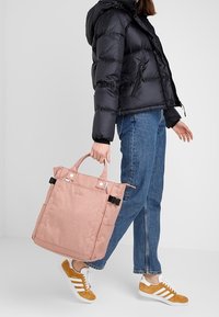 Sac cabas rose en tissu texturé, forme ferme, doubles poignées supérieures et accents noirs. Associé à une doudoune noire et un jean.