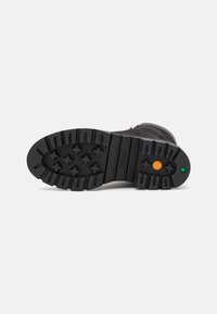 Suela de bota de goma negra con un patrón de tracción robusto, que presenta acentos circulares en naranja y verde. Texturizada para mayor agarre y durabilidad.
