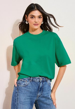 Frau mit langem dunklem Haar, trägt cremefarbene Creolen, ein grünes Kurzarmshirt und blaue Jeans, steht mit einer Hand in der Tasche.