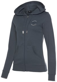 Sudadera con cremallera de color gris oscuro, confeccionada con tejido suave, cuenta con capucha, dos bolsillos frontales y un logotipo circular en el pecho.