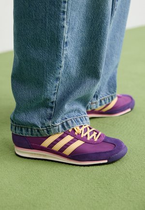 Osoba nosząca fioletowo-beżowe sneakersy Adidas z żółtymi sznurówkami i niebieskimi dżinsami stoi na zielonym dywanie.