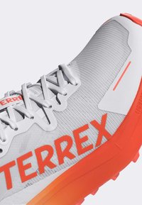 Scarpa da ginnastica leggera con tomaia in rete grigia e dettagli arancioni, superficie testurizzata e un logo "TERREX" ben visibile.