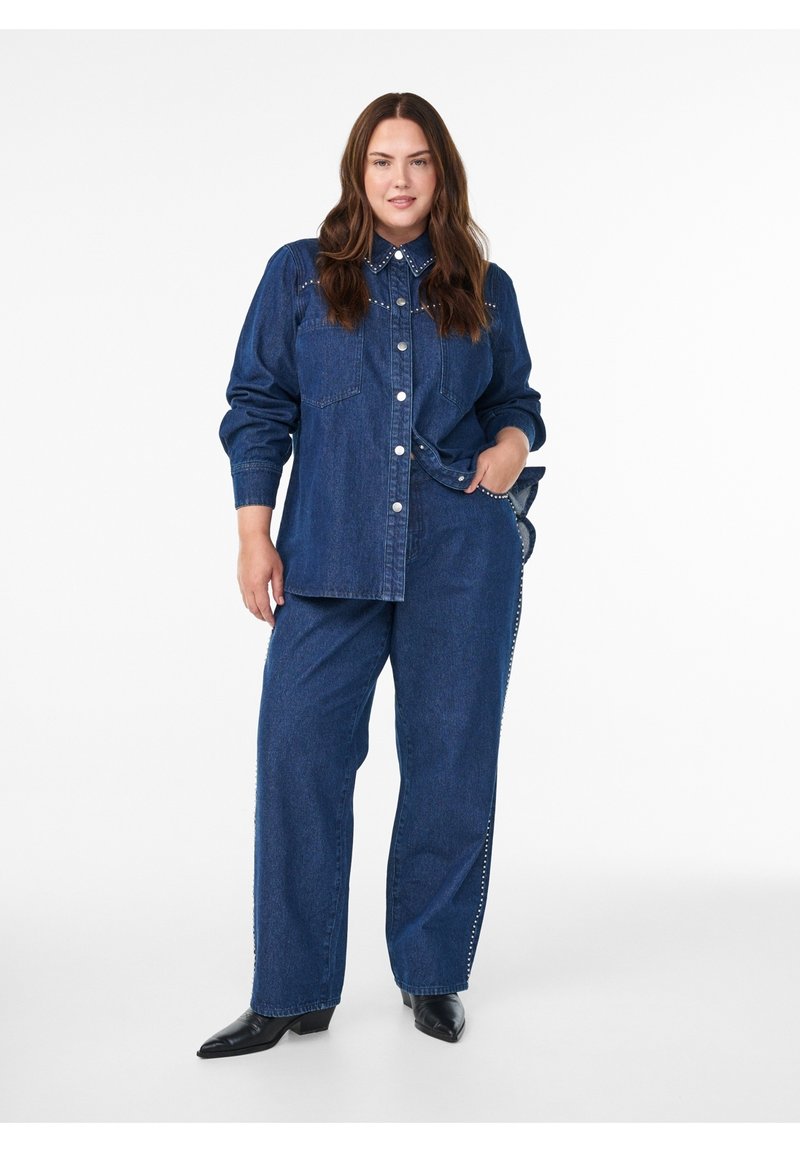 Kvinde med langt brunt hår iført mørkeblå denimskjorte og matchende vidde bukser med sorte spidse sko, stående med den ene hånd i lommen.