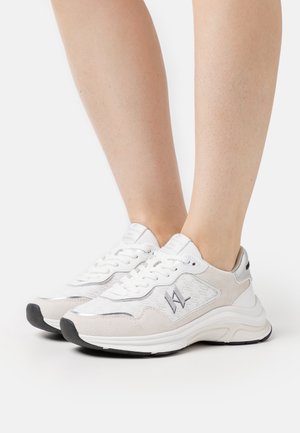 Baskets blanches et beiges à lacets avec semelles noires portées aux pieds, présentant un tissu à motif subtil et un petit logo brodé sur le côté.