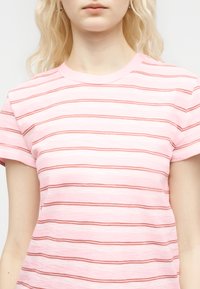 Jeune femme portant un t-shirt à manches courtes rose clair avec de fines rayures horizontales rouges, vue du cou jusqu'à la taille supérieure.