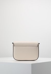 Pieni kermaisen värinen olkalaukku, jossa on sileä nahkapinta, taitettava läppä ja mustat reunat, asetettuna minimalistiselle jalustalle.