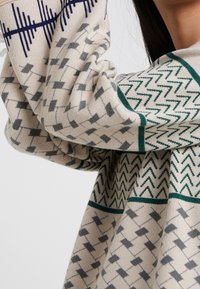 Pull beige avec des motifs géométriques en marine, gris et vert. Présente un tricot texturé et des poignets retroussés, mettant en valeur un design décontracté.