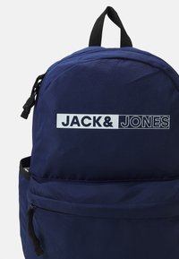 Zaino blu navy con tessuto testurizzato, dotato di un grande etichetta con logo e una tasca frontale con zip. Include una maniglia superiore imbottita per il trasporto.