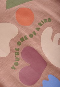 Multikleurige abstracte vormen op roze stof met groene tekst die zegt "JE BENT UNIEK" en om de vormen heen gekruld is.