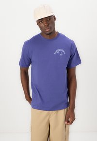 Lilla bomulls t-skjorte med korte ermer, med et hvitt grafisk trykk på brystet. Båret med beige shorts og en lys hette.