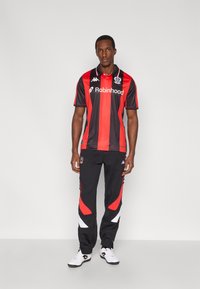 Μαύρη και κόκκινη αθλητική φανέλα με γιακά και λογότυπο, συνδυασμένη με μαύρο παντελόνι φόρμας που διαθέτει κόκκινες και λευκές λεπτομέρειες και φερμουάρ στους αστραγάλους.