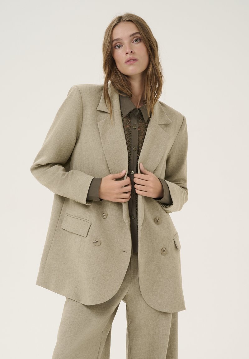 Lys beige dobbeltradet blazer med to forlommer, tekstureret stof og smalere revers, båret over en mørkegrøn skjorte med knapper.