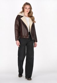Jachetă din piele maro cu guler din blană sintetică, combinată cu pantaloni negri cu picioare ample și pantofi negri, având un fermoar în față și buzunare laterale.