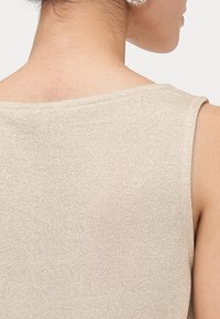 Beige ilmanhihaista puseroa, jossa on hieno, teksturoitu neule. Pehmeä, kiiltävä pinta, jossa on hienovaraisia kimalluksia, pyöreä pääntie ja siistit reunat.