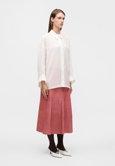 Chemise blanche transparente à boutons avec motif texturé, associée à une jupe midi en suède rose. Collants noirs et escarpins blancs à bout pointu complètent le look.