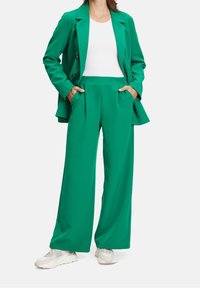 Mujer vistiendo un traje verde con pantalones de pierna ancha y blazer sobre una camiseta blanca, con las manos en los bolsillos, combinando con zapatillas blancas, sobre un fondo liso.