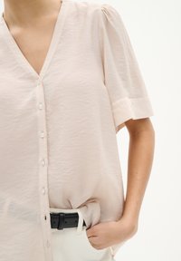 Blusa rosa chiaro a V con maniche corte, caratterizzata da una chiusura con bottoni e un tessuto morbido e strutturato. Indossata infilata in pantaloni bianchi.