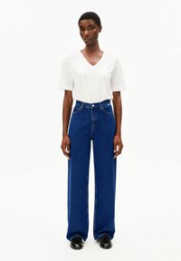 Weißes T-Shirt mit V-Ausschnitt, kombiniert mit hoch taillierter, weit geschnittener blauer Denim-Jeans. Die Jeans hat eine glatte Textur und leichte Used-Details. Schwarze Schuhe vervollständigen das Outfit.