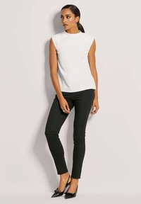 Weißes ärmelloses Oberteil mit Rundhalsausschnitt und lockerer Passform, kombiniert mit eng anliegenden schwarzen Jeans und spitzen schwarzen Pumps. Glatte Stofftextur.