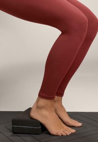 Bordové legíny priliehajúce na telo, skombinované s bosými nohami na čiernom obdĺžnikovom bloku. Povrchová textúra vyzerá hladko so slabým leskom.