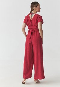 Jumpsuit rosso, texture a coste, con maniche corte e gambe larghe. Fiocco in vita sul retro, apertura a chiave sul collo. Indossato con tacchi di colore chiaro.