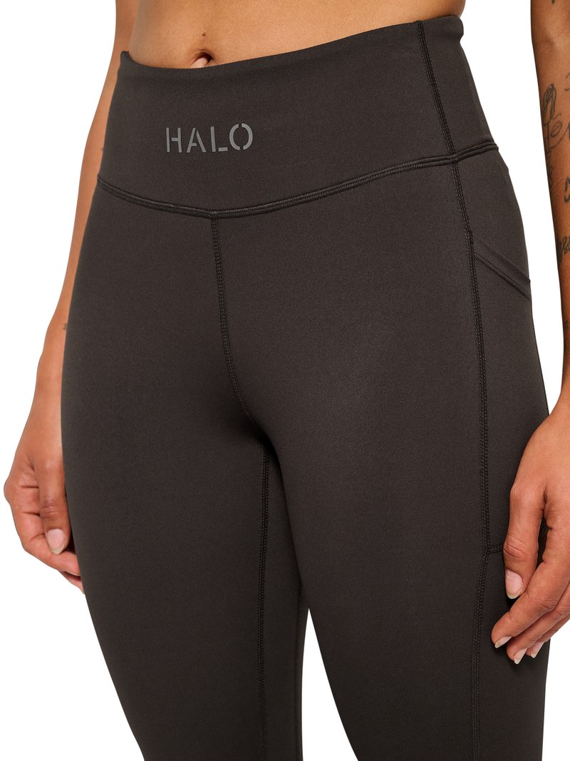Zbliżenie na środkową część ciała w czarnych legginsach z wysokim stanem z logo "HALO", widocznym przeszyciem szwu oraz boczną kieszenią na prawej nodze.