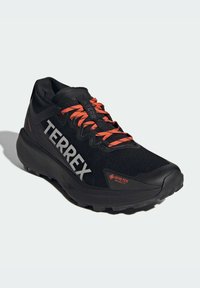 Chaussures de sport noires avec tige en textile, arborant des lacets orange vif, un logo "TERREX" bien visible et une semelle extérieure en caoutchouc texturé.