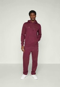 Completo in felpa bordeaux con cappuccio e pantaloni tuta, dotato di tasca a marsupio, cappuccio con stringa e logo ricamato. Tessuto liscio e vestibilità comoda.