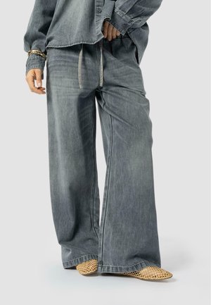 Persona que lleva pantalones de denim gris holgados con cintura de cordón, combinados con una camisa de denim gris a juego y zapatos puntiagudos de mimbre beige.