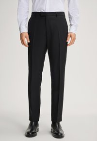 Schwarze Hose mit glatter Textur, mit Falten und tailliertem Bund, kombiniert mit schwarzen eleganten Schuhen und einem weißen Hemd mit Punkten.