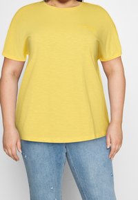 T-shirt giallo a maniche corte realizzato in tessuto morbido, con scollatura rotonda e un piccolo logo sul petto. Indossato con jeans chiari.