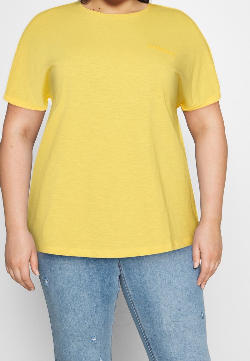 T-shirt giallo a maniche corte realizzato in tessuto morbido, con scollatura rotonda e un piccolo logo sul petto. Indossato con jeans chiari.