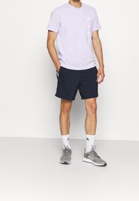 Camiseta de manga corta morada clara con tres logos blancos de Adidas en el lado izquierdo, combinada con pantalones cortos navy y zapatillas deportivas grises con suelas blancas.