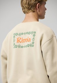 Beige Sweatshirt mit einem floralen Rahmen um die Grafik und dem Wort "Rivvia" in Orange. Rundhalsausschnitt und lässige Passform mit sichtbaren Nahtdetails.