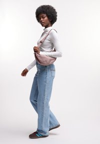 Borsa a spalla marrone realizzata in tessuto liscio, forma curva, cinturino regolabile, abbinata a jeans azzurri chiari e un dolcevita bianco.