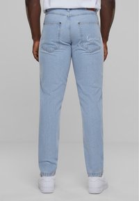 Jeans in denim azzurro chiaro con taglio dritto, dotati di due tasche posteriori e dettagli del logo cucito sulla tasca destra, abbinati a sneaker bianche.