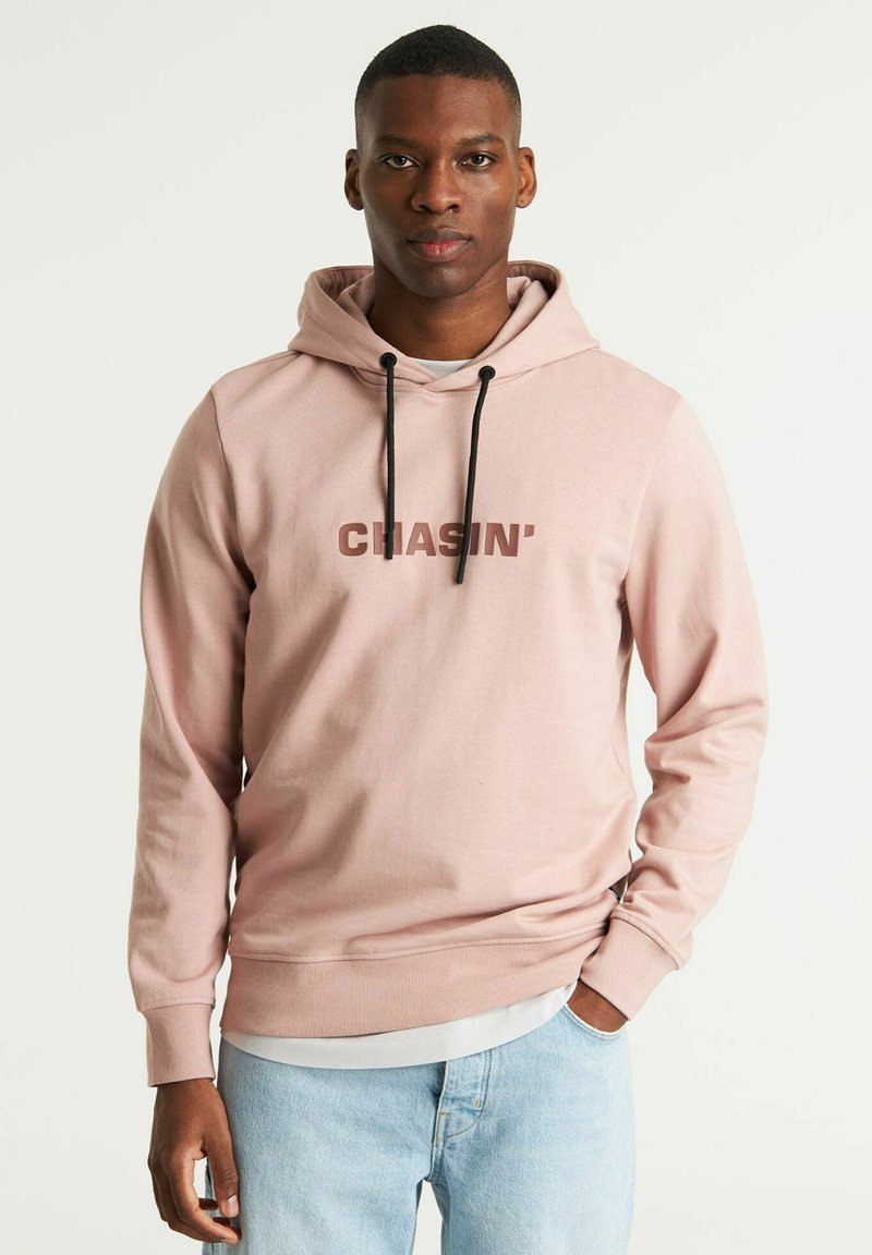 Felpa rosa chiaro con stampa frontale "CHASIN," cappucci neri e vestibilità rilassata; abbinata a jeans azzurri.