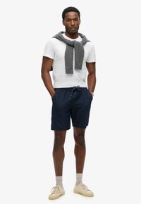 Shorts bleu marine à pois avec une taille élastique, associés à un t-shirt blanc et un pull gris drapé sur les épaules. Baskets beiges aux pieds.