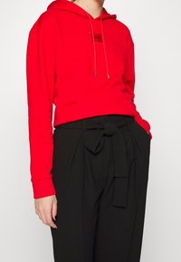 Červená mikina s kapucí a přední kapsou, doplněná o značkovou nášivku, v kombinaci s černými kalhotami s vysokým pasem a uzlem na pásku.