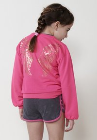 Sweatshirt rose avec des motifs d'ailes dorées métalliques dans le dos, associé à un short gris foncé bordé d'une finition rose et de détails en forme d'étoiles.