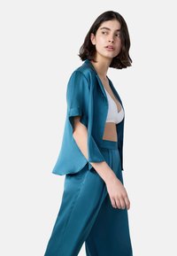 Junge Frau mit kurzem dunklem Haar trägt ein teal-farbenes Satinoberteil und eine -hose über einem weißen Bralette und blickt nach links vor einem schlichten Hintergrund.