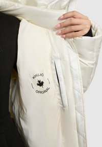 Cremefarvet pufferjakke indvendig med en quiltet struktur, prydet med en sort logo-print og en sidelomme til opbevaring.