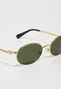 Gafas de sol de metal dorado con lentes ovalados verdes. Presentan una patilla texturizada con acentos de cristal y un brazo de carey.
