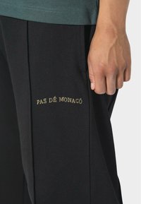 Czarne spodnie z miękkiego materiału, z plisami i haftowanym detalem "PAS DE MONACO" w kolorze złotym na lewej nodze.