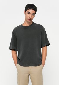 Young man with short black hair wearing a loose dark gray t-shirt and beige pants with hands in pockets, against a plain light background.