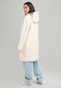 Beige raincoat with a hood, featuring a straight cut, snap-button cuffs, and a smooth, water-resistant texture, paired with light blue baggy jeans.