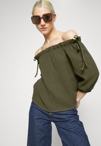 Top verde oliva sem mangas com decote com babados e laços, tecido texturizado, combinado com jeans azuis de perna larga e óculos de sol castanhos de tamanho exagerado.