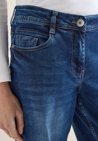 Gros plan sur une personne portant un jean en denim bleu et une chemise blanche à manches longues, la main reposant le long du corps.